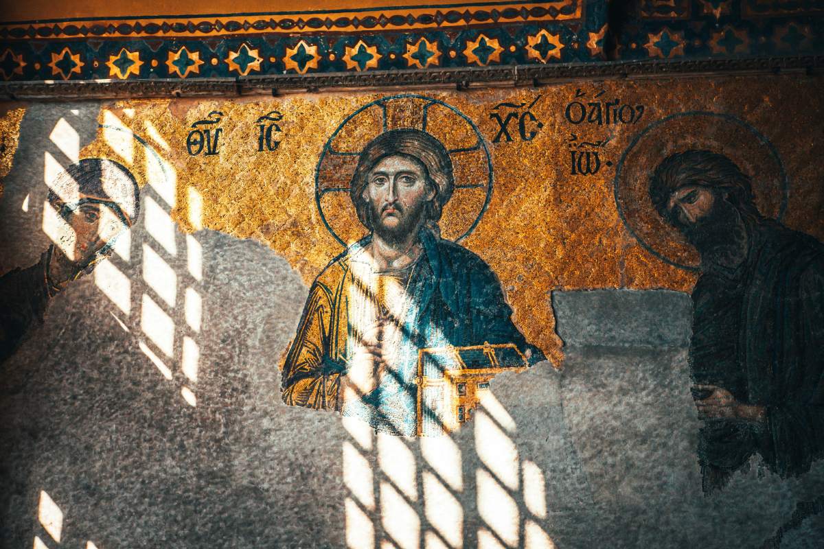 A Byzantine mosaic of Christ with a golden background, partially worn, illuminated by sunlight casting shadows through a lattice window.